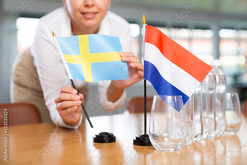 Unrecognizable woman preparing room for international negotiations and communication discussions of leaders. Lady sets miniatures flags of Netherlands and Sweden on table. Unfocused shot