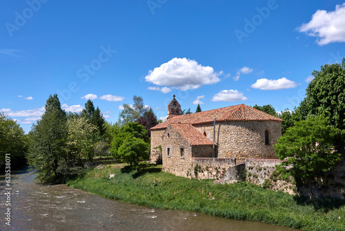 Landscape with a nice old church built with pebbles, in the middle of green trees and next to a river, under a blue sky and some clouds © Sylvain