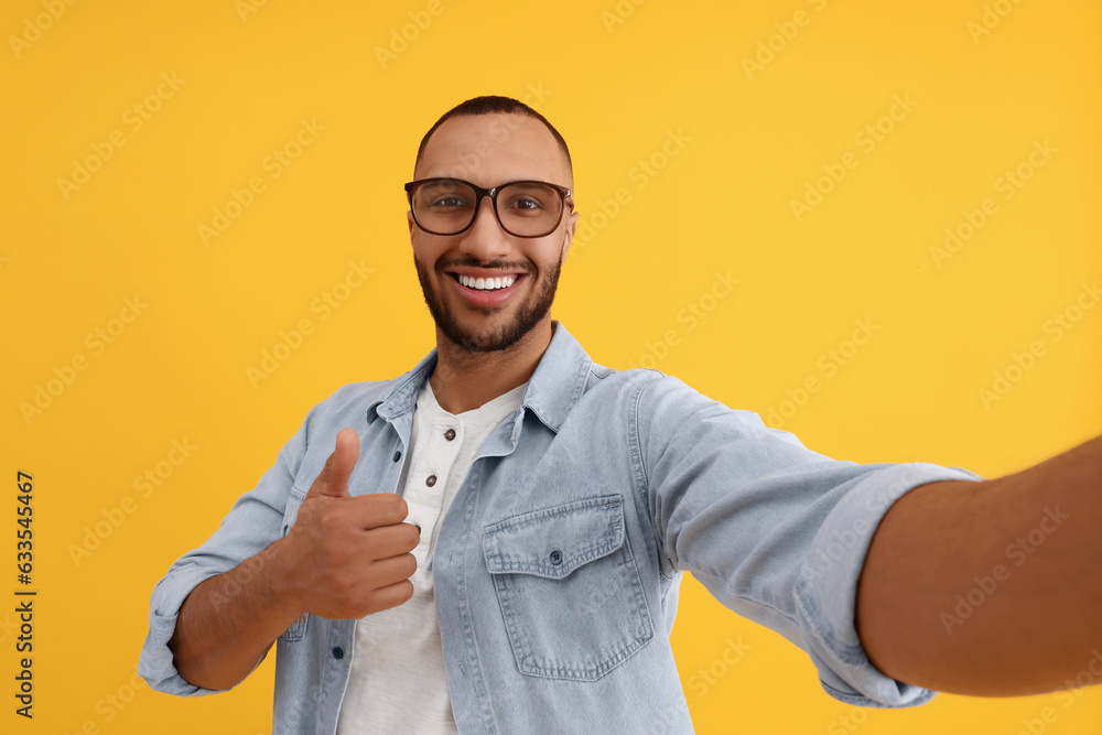 Smiling young man taking selfie and showing thumbs up on yellow background Smiling young man taking selfie and showing thumbs up on yellow background