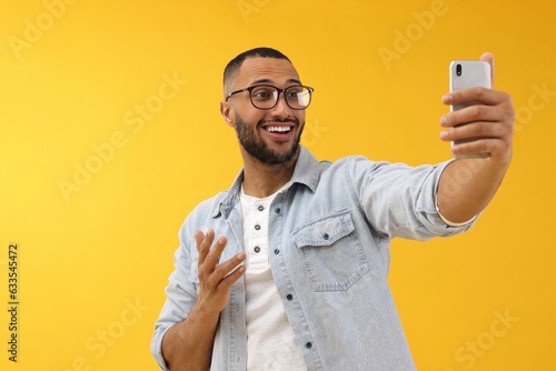 Smiling young man taking selfie with smartphone on yellow background © New Africa