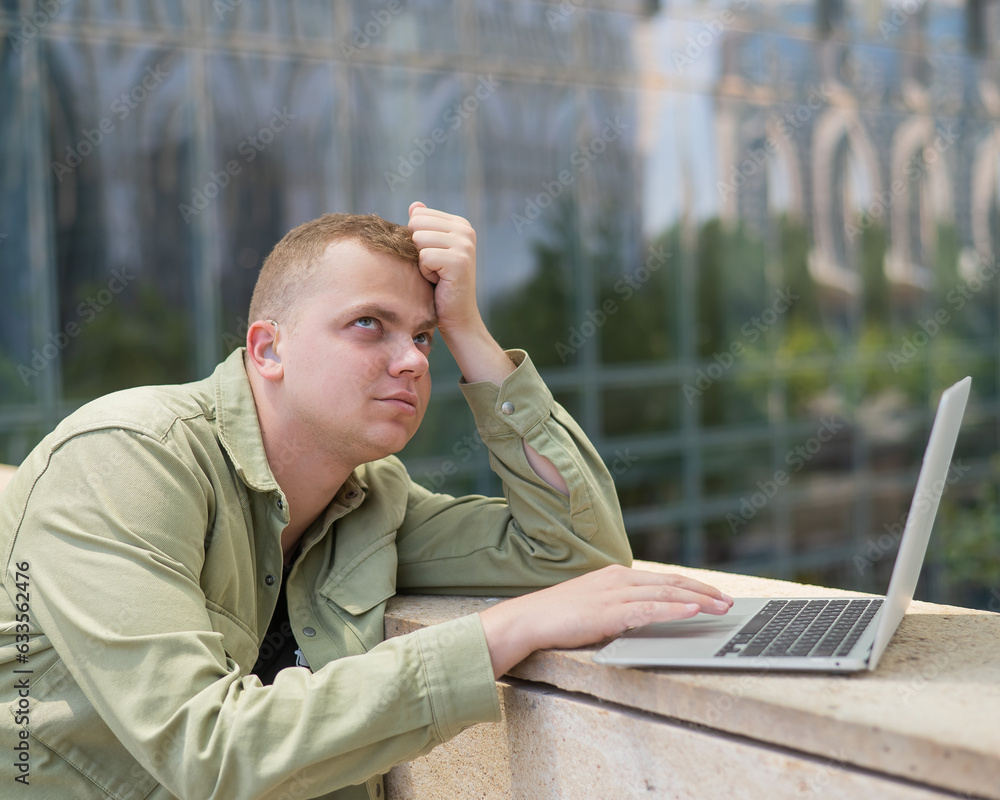 Sad guy with hearing aid using laptop outdoors. Sad guy with hearing aid using laptop outdoors.