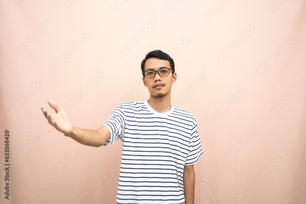 Asian man in glasses wearing casual striped t-shirt, greeting pose and smiling. Isolated beige background. Asian man in glasses wearing casual striped t-shirt, greeting pose and smiling. Isolated beige background.