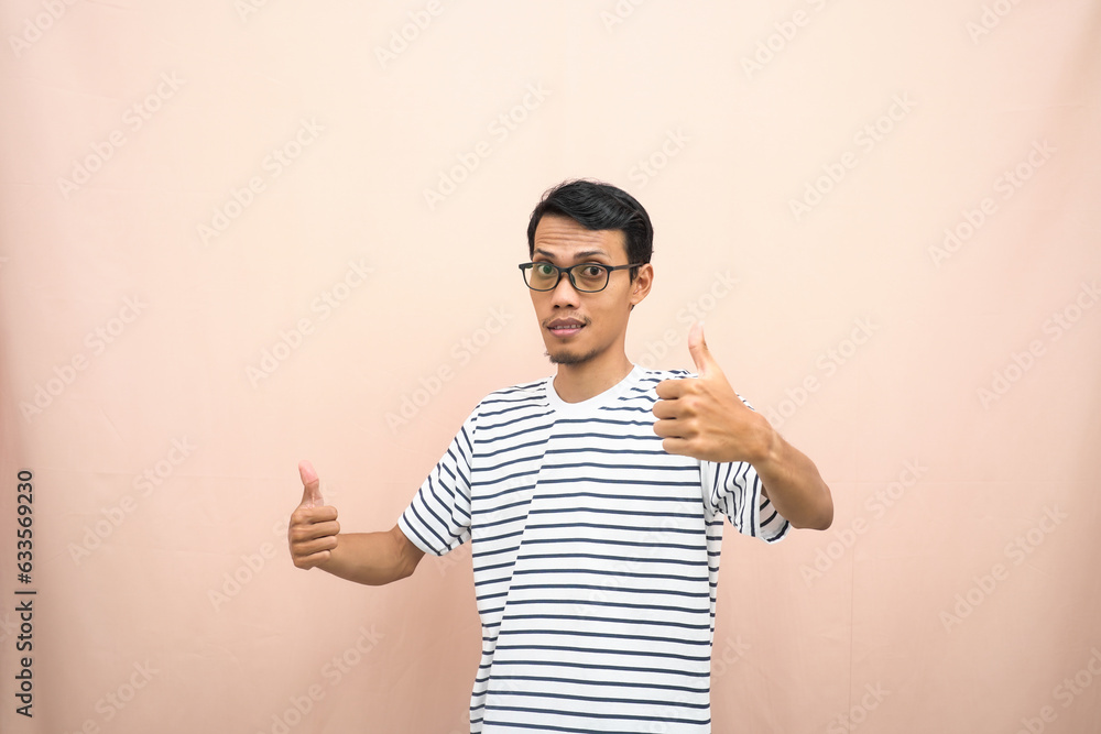 Asian man in glasses wearing casual striped shirt, posing giving thumbs up gesture or okay that agree. Isolated beige background. Asian man in glasses wearing casual striped shirt, posing giving thumbs up gesture or okay that agree. Isolated beige background.