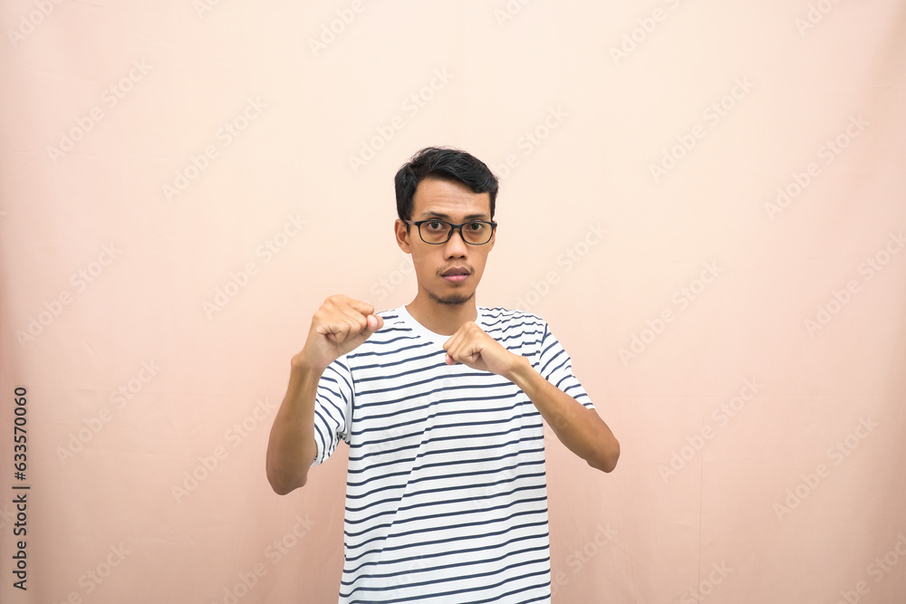 Asian man with glasses wearing casual striped shirt, fist fist pose, indicating strong or ready to fight. Isolated beige background. Asian man with glasses wearing casual striped shirt, fist fist pose, indicating strong or ready to fight. Isolated beige background.