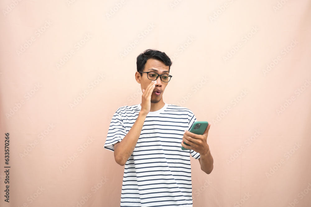 Asian man in glasses wearing casual striped t-shirt, whispering pose giving good news and surprise while holding smartphone. Isolated beige background. Asian man in glasses wearing casual striped t-shirt, whispering pose giving good news and surprise while holding smartphone. Isolated beige background.