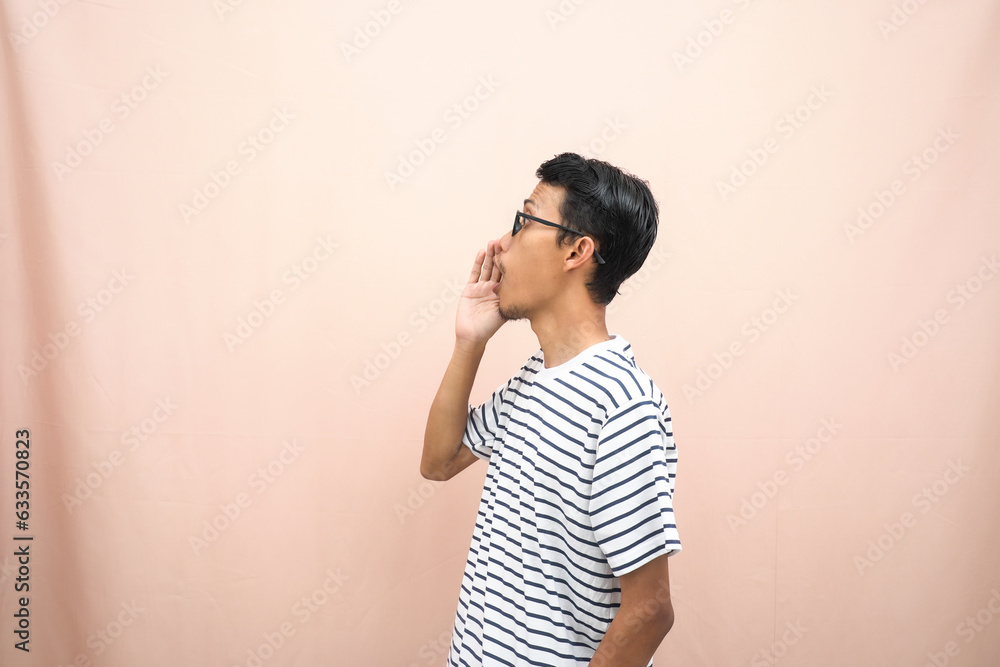 Asian man in glasses wearing casual striped shirt, shouting pose with hand near mouth, giving information. Isolated beige background. Asian man in glasses wearing casual striped shirt, shouting pose with hand near mouth, giving information. Isolated beige background.