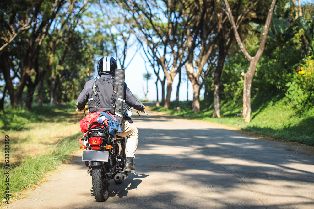 Back view of motorcyclist in black jacket and helmet riding motorbike along country road between tall green trees. Back view of motorcyclist in black jacket and helmet riding motorbike along country road between tall green trees.
