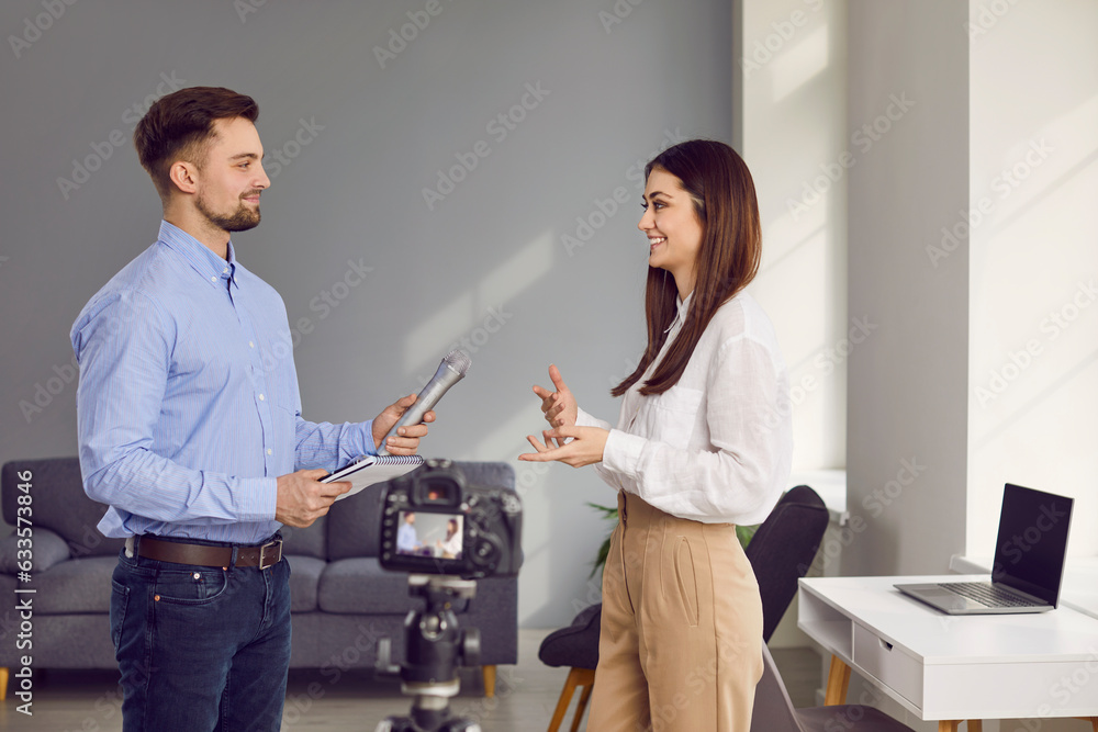 Fototapeta premium Professional cheerful male journalist standing with microphone interviewing young businesswoman in office. Portrait of successful young woman giving interview to reporter at workplace