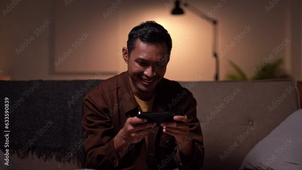 Asian Man playing online game with mobile smartphone sitting on a couch in the living room at home. Asian Man playing online game with mobile smartphone sitting on a couch in the living room at home.