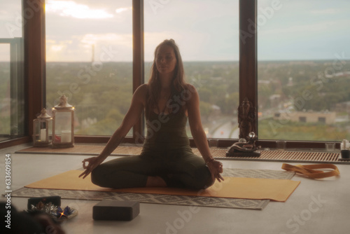 Woman in green sports attire practicing yoga and meditation in lotus pose against a panoramic cityscape backdrop in an apartment