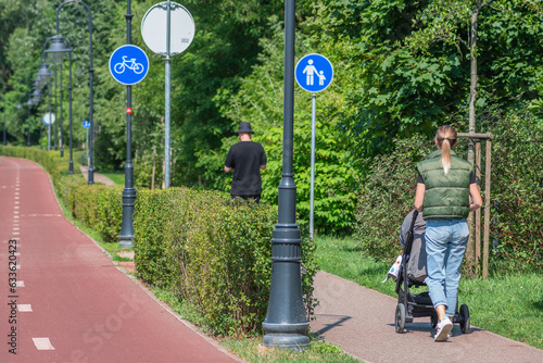 Young blonde hair girl or mother with baby carriage, pram or stroller walking near the bike road with blue road sign or signal of bicycle lane among green trees