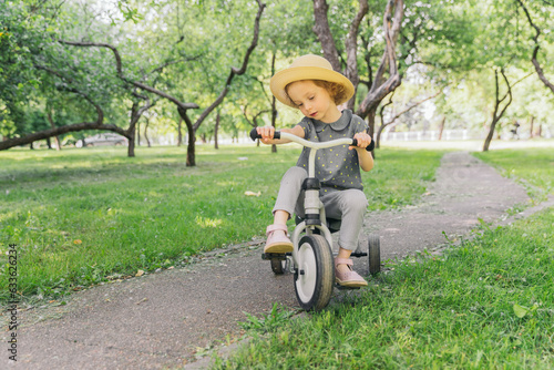 A little girl rides along a path in an apple orchard. The girl is wearing a straw hat. The weather is fine outside, the sun is shining. The frame is dynamic. The baby turns the steering wheel.