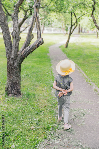 A little girl walks along the path in the apple orchard. The girl is wearing a straw hat. The weather is fine outside, the sun is shining.