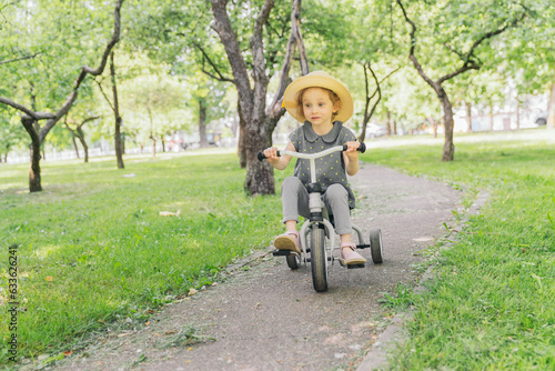 A little girl rides along a path in an apple orchard. The girl is wearing a straw hat. The weather is fine outside, the sun is shining. The frame is dynamic. The frame is dynamic.