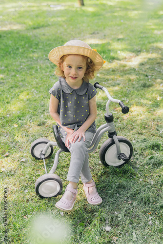 The baby girl is sitting on a bicycle. The tricycle is in the apple orchard. The girl is wearing a straw hat. The girl smiles. The photo was taken on a clear summer day.