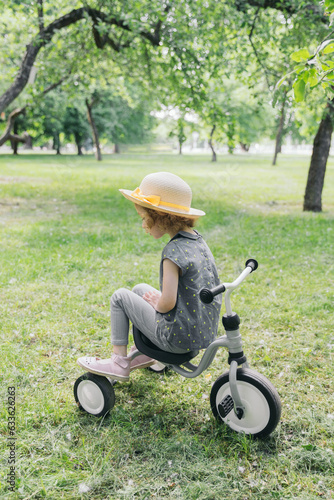 Baby girl on a bicycle in an apple orchard. In the foreground is a girl on a bicycle, back seated, and in the background are apple trees in a blur. The girl is dressed in a straw hat.