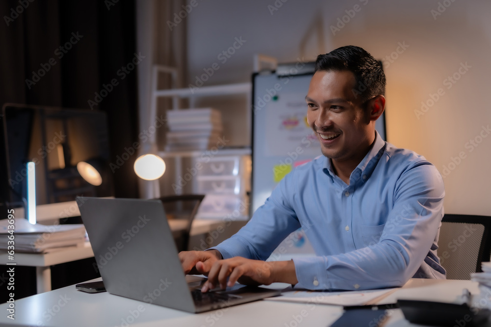 Commitment Asian businessman sitting at desk in office Working overtime late at night with document and laptop computer. Overtime work concept.