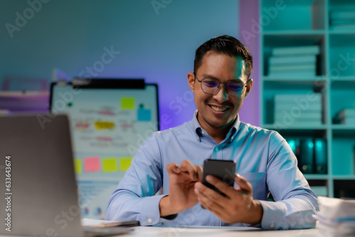 Committed Asian businessman is using the smartphone and working overtime late at night in the office.