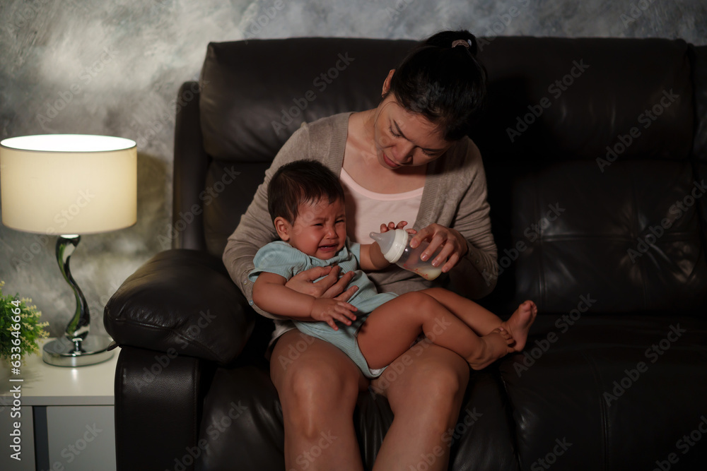 stressed mother trying to feeding milk bottle to crying infant baby on sofa in the living room at night stressed mother trying to feeding milk bottle to crying infant baby on sofa in the living room at night