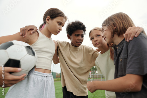 Football team of four cute intercultural schoolchildren consulting about game while standing in front of camera at break after play