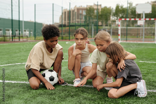Four schoolchildren in activewear standing on knees on green football field or sports ground and having break between outdoor trainings