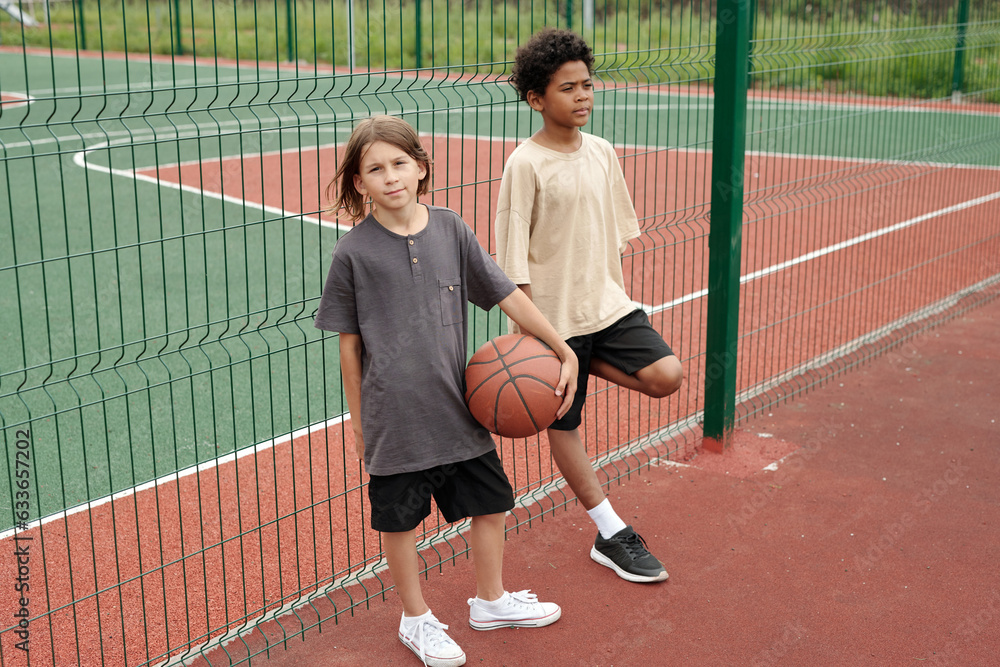 Cute schoolboy with ball for playing basketball looking at camera while standing by green metallic fence next to African American buddy