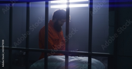 Believing male prisoner in orange uniform kneels near the bed, prays to God in prison cell holding Bible. Criminal serves imprisonment term for crime in jail or detention center. Faith in God concept.