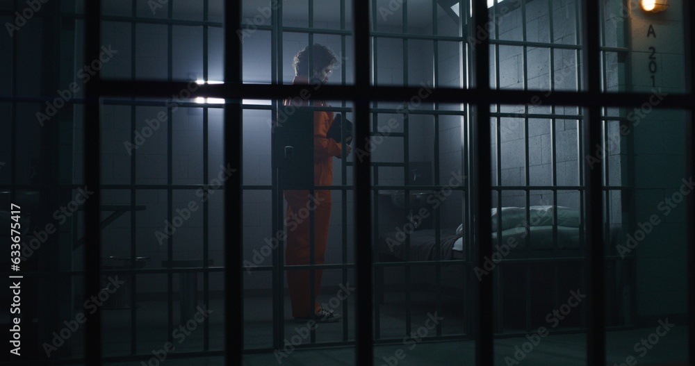 Prisoner in orange uniform kneels near bed, prays to God in prison cell holding Bible. Jailer walks, watches criminal. Inmate serves imprisonment term in jail. View through metal bars. Faith in God.