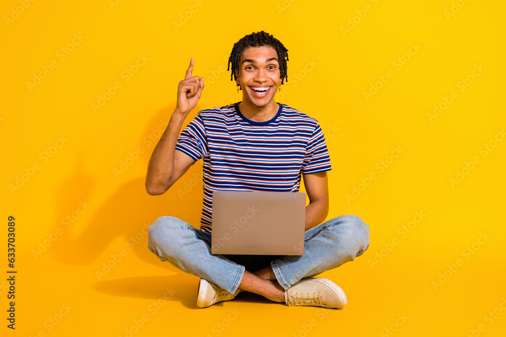Full length portrait of overjoyed handsome guy sit floor use netbook point finger up empty space isolated on yellow color background Full length portrait of overjoyed handsome guy sit floor use netbook point finger up empty space isolated on yellow color background