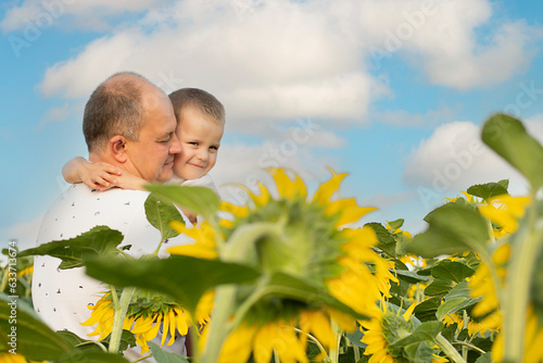 Father's day concept. A little boy in his father's arms gently hugs and cuddles, standing in a field with yellow sunflowers against the background of a blue sky. A symbol of peace. Symbol of Ukraine.