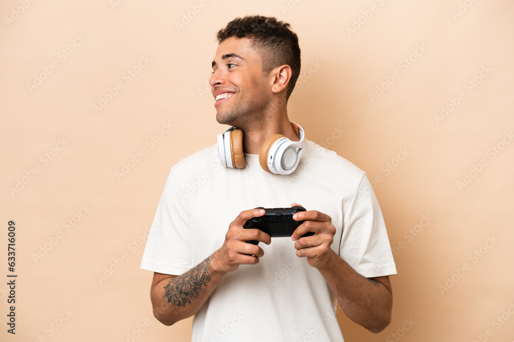 Young Brazilian man playing with a video game controller isolated on beige background looking side Young Brazilian man playing with a video game controller isolated on beige background looking side
