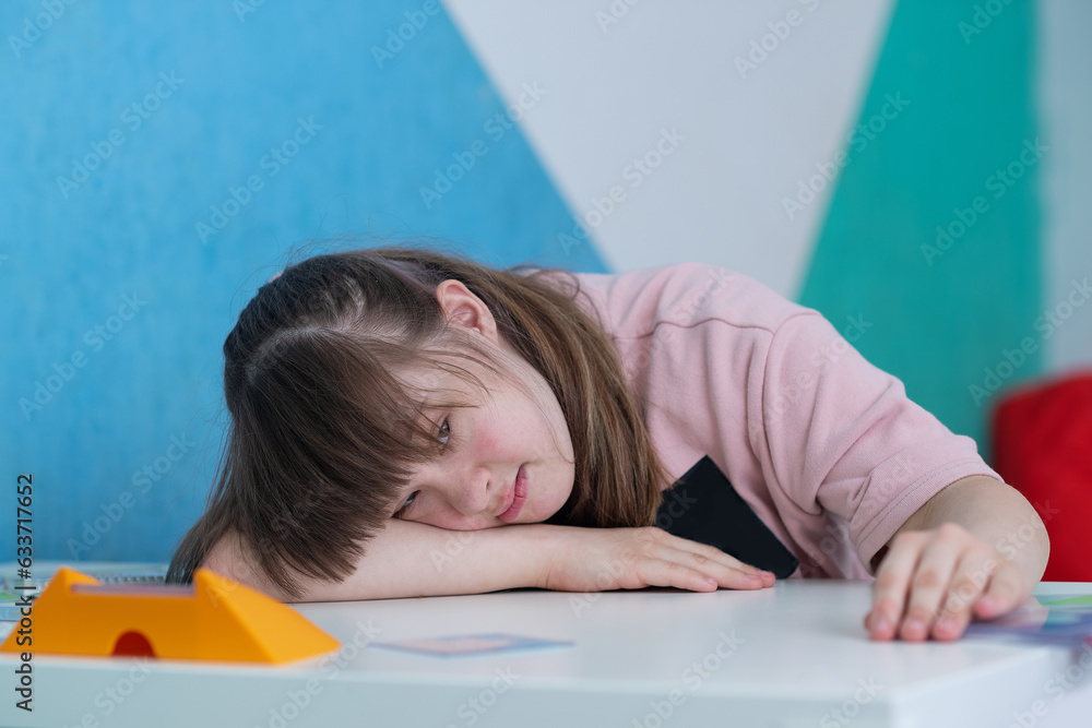 Sunny girl lies with his head on his folded hands on the table in the educational room. World day of people with down syndrome. A special child Sunny girl lies with his head on his folded hands on the table in the educational room. World day of people with down syndrome. A special child
