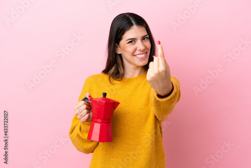 Young Italian woman holding a coffee maker isolated on pink background doing coming gesture