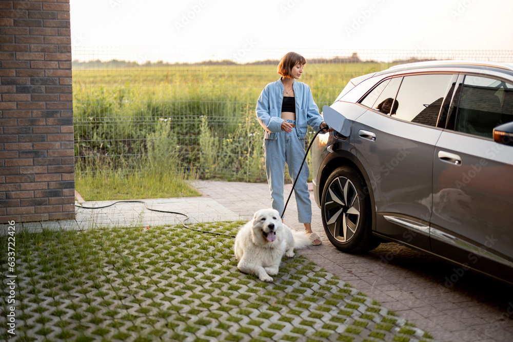 Woman charges electric car, standing with her cute white dog near her house on sunset. Concept of green energy, sustainability and modern lifestyle
