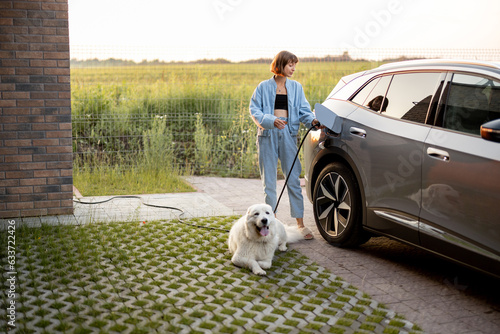 Obraz na płótnie Woman charges electric car, standing with her cute white dog near her house on sunset. Concept of green energy, sustainability and modern lifestyle w salonie