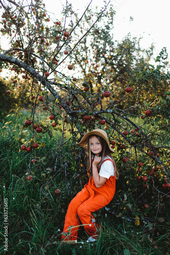 A girl in a hat is sitting on a branch of an apple tree full of red apples