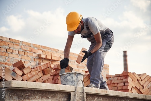 Bucket on the ground, holding a brick. Handsome Indian man is on the construction site