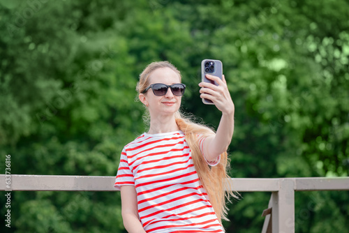 Blonde girl in sunglasses and T-shirt takes a selfie or shoots a video on the smartphone background of green foliage of trees.