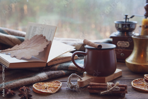 Autumn mood, autumn atmosphere. A cup of hot coffee, a plaid blanket, a coffee mill, cinnamon sticks, star anise, a book on a wooden windowsill on a rainy day.