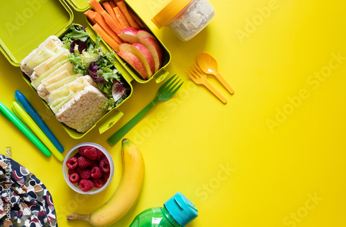 School lunch box with sandwiches, carrot sticks, apple, banana, lettuce, hummus and raspberries. Healthy school lunch concept. Yellow background. Top view. Place for text.