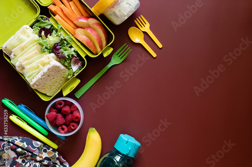 School lunch box with sandwiches, carrot sticks, apple, banana, lettuce, hummus and raspberries. Healthy school lunch concept. Burgundy background. Top view. Place for text