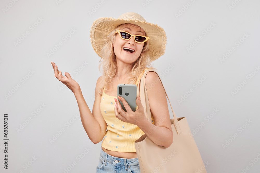Portrait positive mature blond woman in straw hat, yellow top and sunglasses holding smartphone and shopping bag isolated on white studio background Portrait positive mature blond woman in straw hat, yellow top and sunglasses holding smartphone and shopping bag isolated on white studio background
