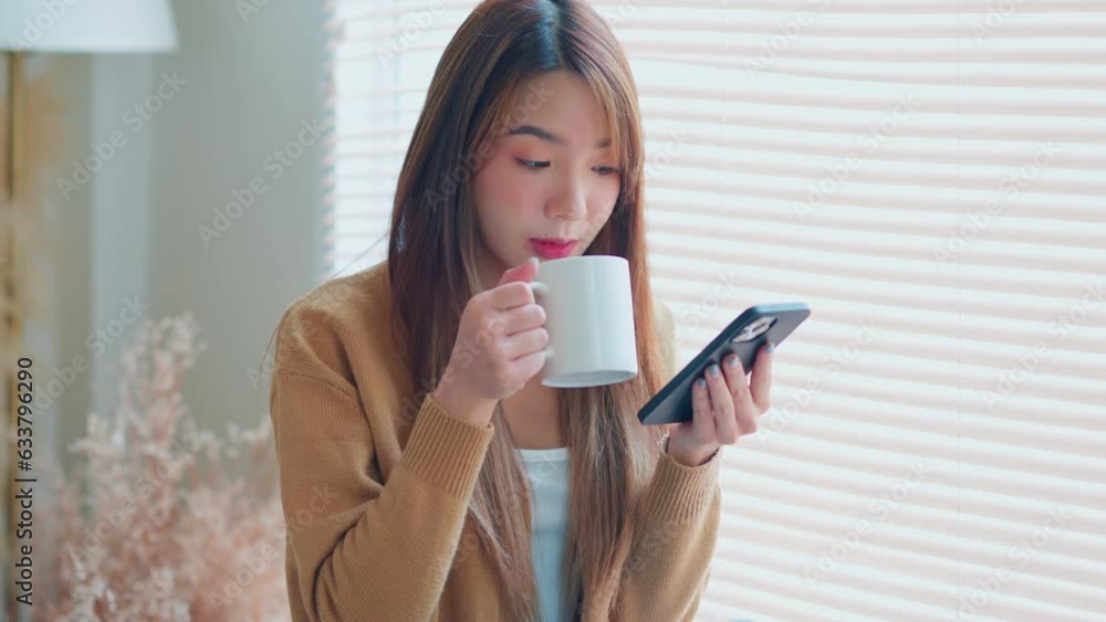 Relaxed young asian woman using mobile smartphone and drinking coffee from a mug while sitting by the window at home, The concept of balance and tranquility. Relaxed young asian woman using mobile smartphone and drinking coffee from a mug while sitting by the window at home, The concept of balance and tranquility.