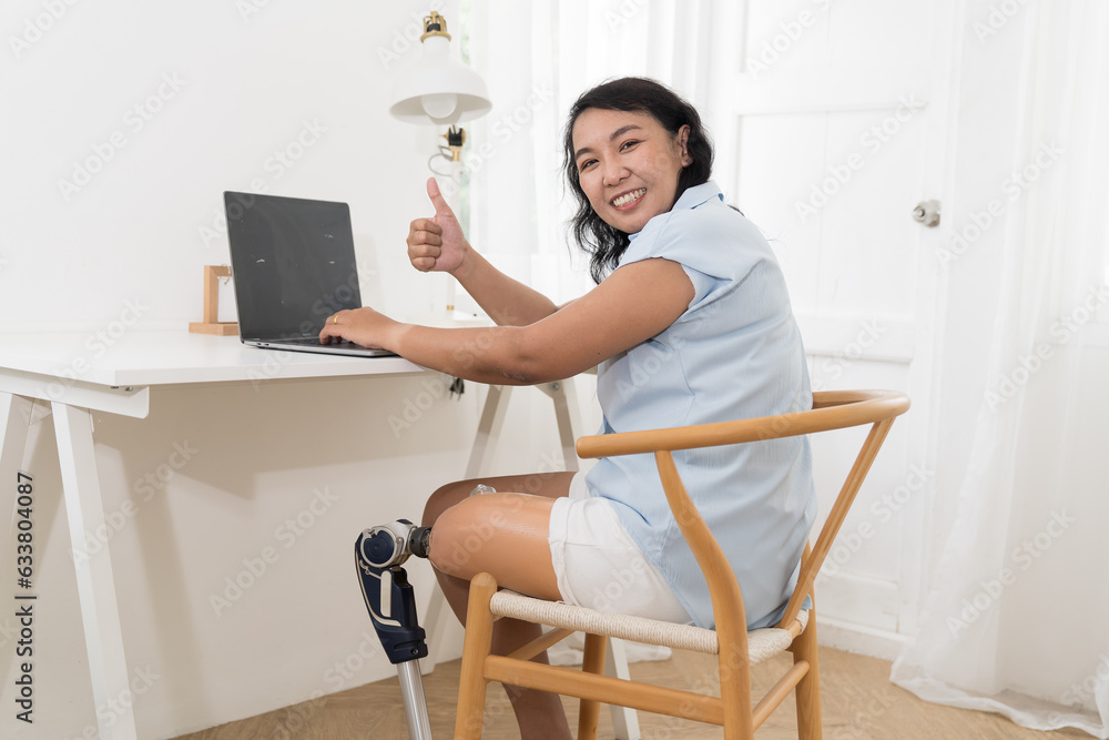 Happy smiling Asian female with prosthetic leg sitting and working at home. Woman with prosthetic leg. Asian woman with leg prosthesis equipment Happy smiling Asian female with prosthetic leg sitting and working at home. Woman with prosthetic leg. Asian woman with leg prosthesis equipment