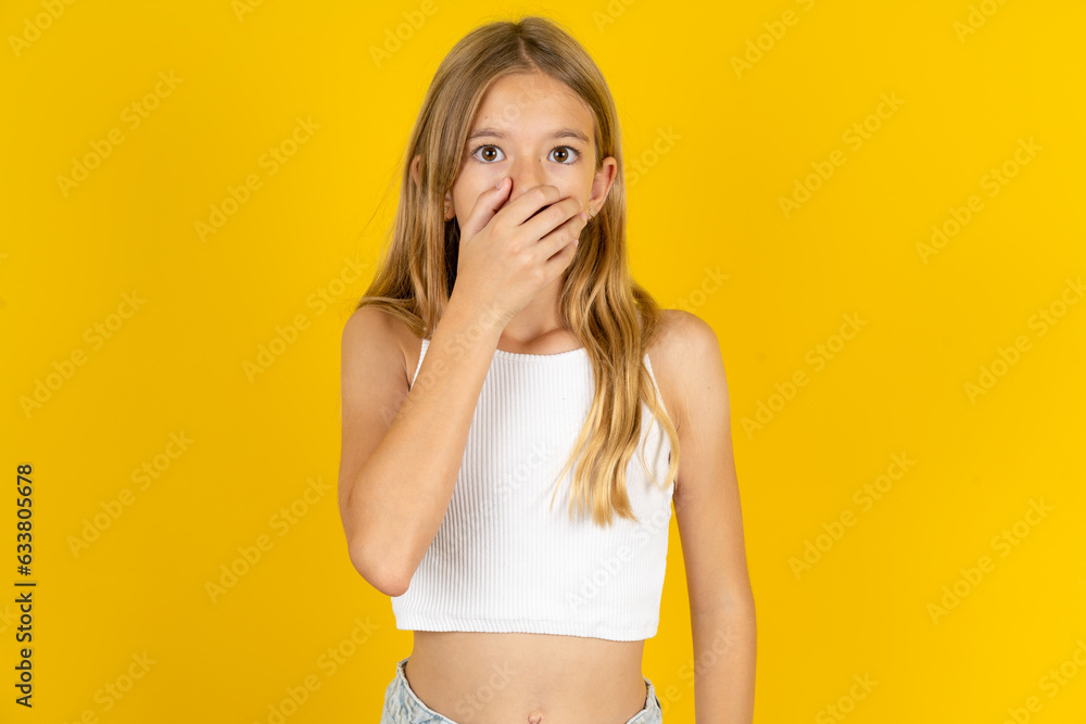 Fototapeta premium Emotional blonde kid girl wearing white T-shirt over yellow studio background gasps from astonishment, covers opened mouth with palm, looks shocked at camera.