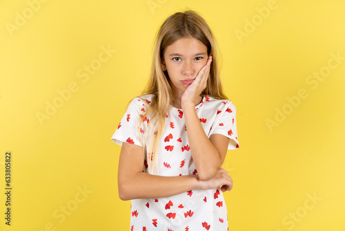 Very bored blonde kid girl wearing polka dot shirt over yellow studio background holding hand on cheek while support it with another crossed hand, looking tired and sick,