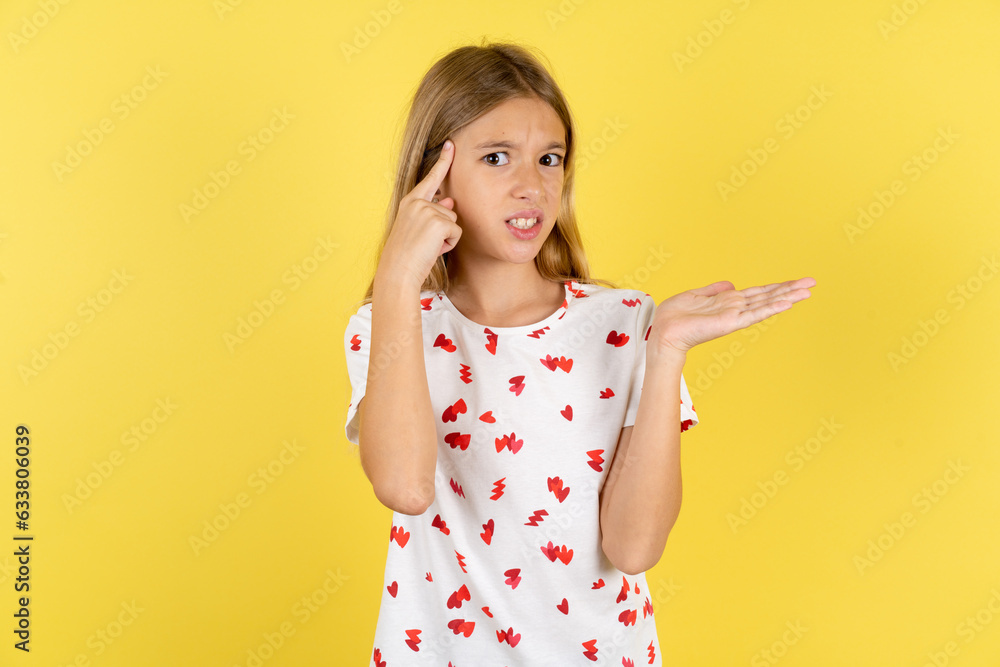 blonde kid girl wearing polka dot shirt over yellow studio background confused and annoyed with open palm showing copy space and pointing finger to forehead. Think about it. blonde kid girl wearing polka dot shirt over yellow studio background confused and annoyed with open palm showing copy space and pointing finger to forehead. Think about it.