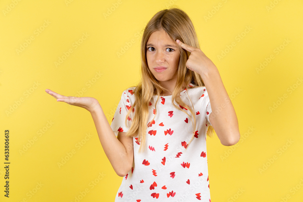blonde kid girl wearing polka dot shirt over yellow studio background confused and annoyed with open palm showing copy space and pointing finger to forehead. Think about it.
