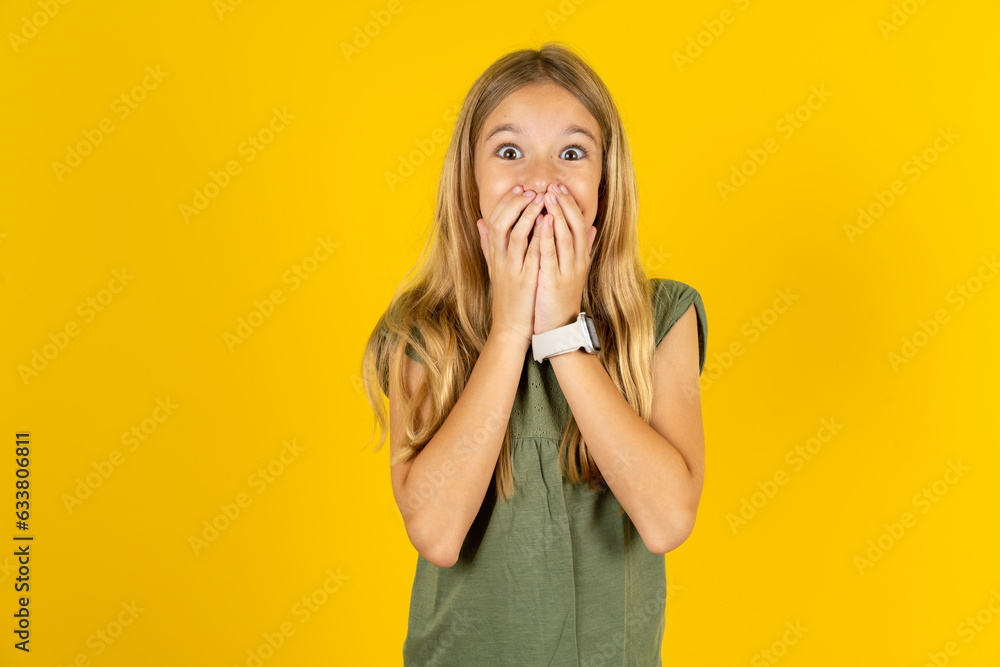 Vivacious blonde kid girl wearing green T-shirt over yellow studio background , giggles joyfully, covers mouth, has natural laughter, hears positive story or funny anecdote - obrazy, fototapety, plakaty 