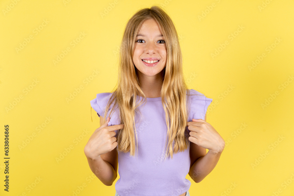 blonde kid girl wearing violet T-shirt over yellow studio background points at his body, being in good mood after going shopping and making successful purchases blonde kid girl wearing violet T-shirt over yellow studio background points at his body, being in good mood after going shopping and making successful purchases